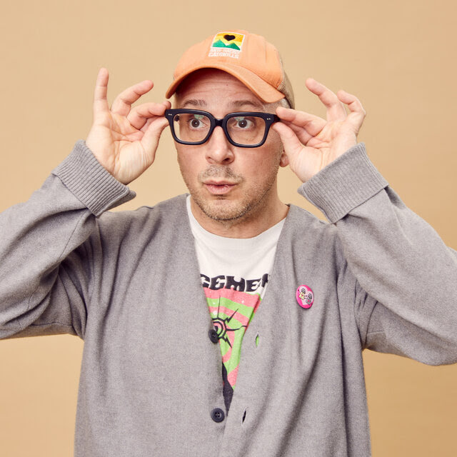 Steve Burns, wearing an orange baseball hat, black-rimmed glasses, and a gray cardigan over a graphic T-shirt.