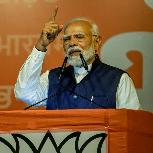 Narendra Modi speaking from behind a lectern. His right arm is raised with his index finger pointing skyward.