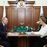 Vladimir Putin and Maria Lvova-Belova sit opposite each other at a table beside a wall with wooden paneling.