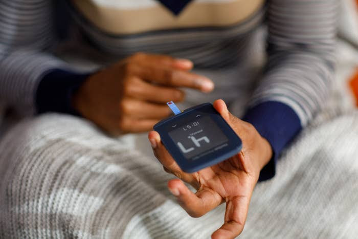 A person holds a glucose meter, indicating a focus on checking blood sugar levels. They are sitting on a knitted blanket