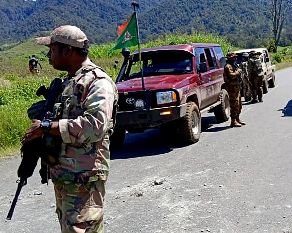 Soldiers holding rifles patrol a road where several trucks are parked.