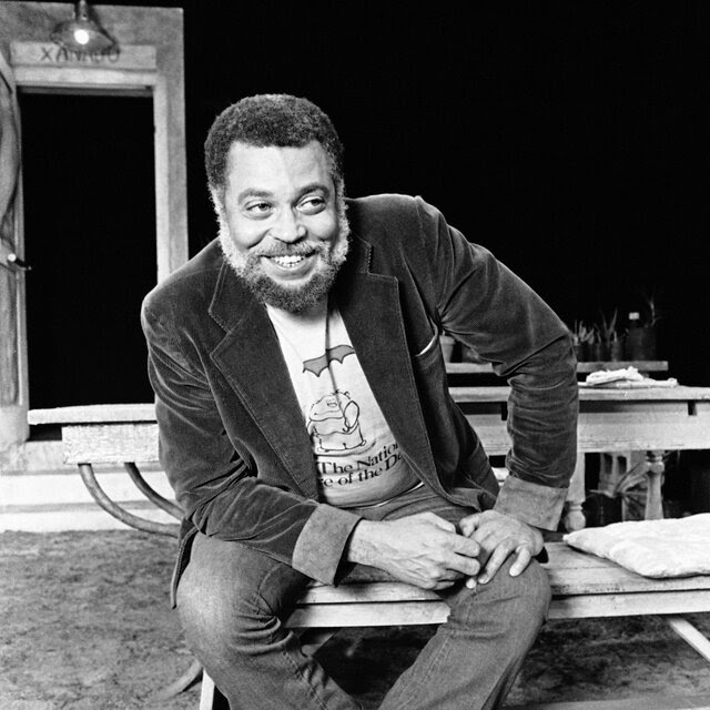 A black and white portrait of a young James Earl Jones sitting on a wooden bench and leaning forward and smiling with his hands on his left knee.