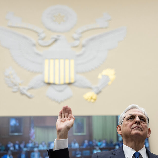 Attorney General Merrick B. Garland, seen from the neck up, holding up his right hand for an oath. A large eagle can be seen on the wall behind him as part of a government logo.