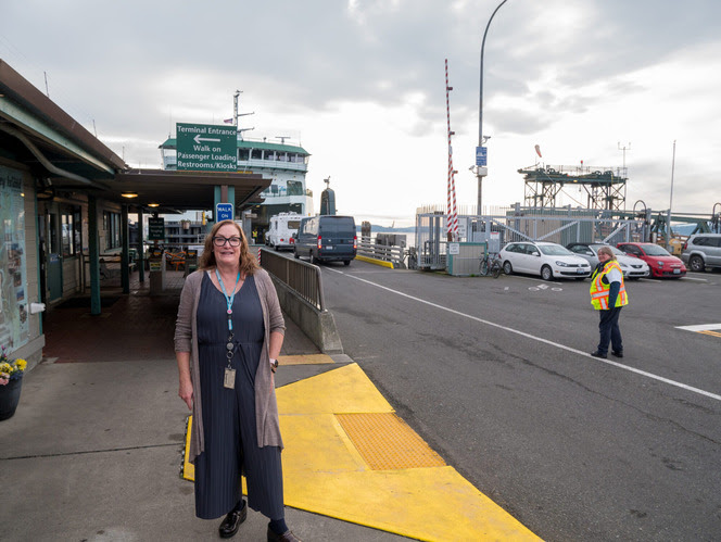 A woman stands near the entrance to the Port Townsend terminal building with parked vehicles and a distant ferry in the background