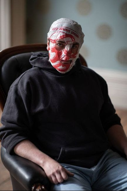 Blindboy Boatclub, with his trademark shopping-bag mask, sits on an armchair in a black hoodie and blue jeans, against a blurry background. He stares at the camera with sullen intensity.