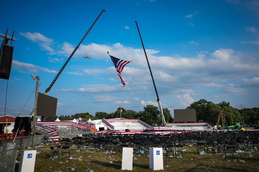 An empty rally venue, with chairs and litter. A huge American flag is held in the sky by two cranes, and a helicopter is flying in the distance.