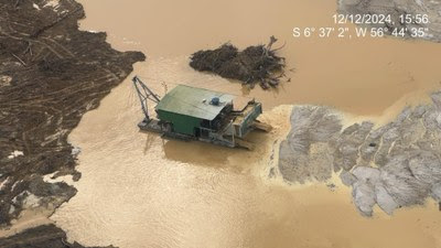 A imagem mostra uma draga flutuante verde operando em um corpo d'água de cor amarelada, cercado por áreas desmatadas e terreno exposto com marcas de erosão. À esquerda, há vegetação removida e troncos no chão. No canto superior direito, a data aparece sobreposta: 