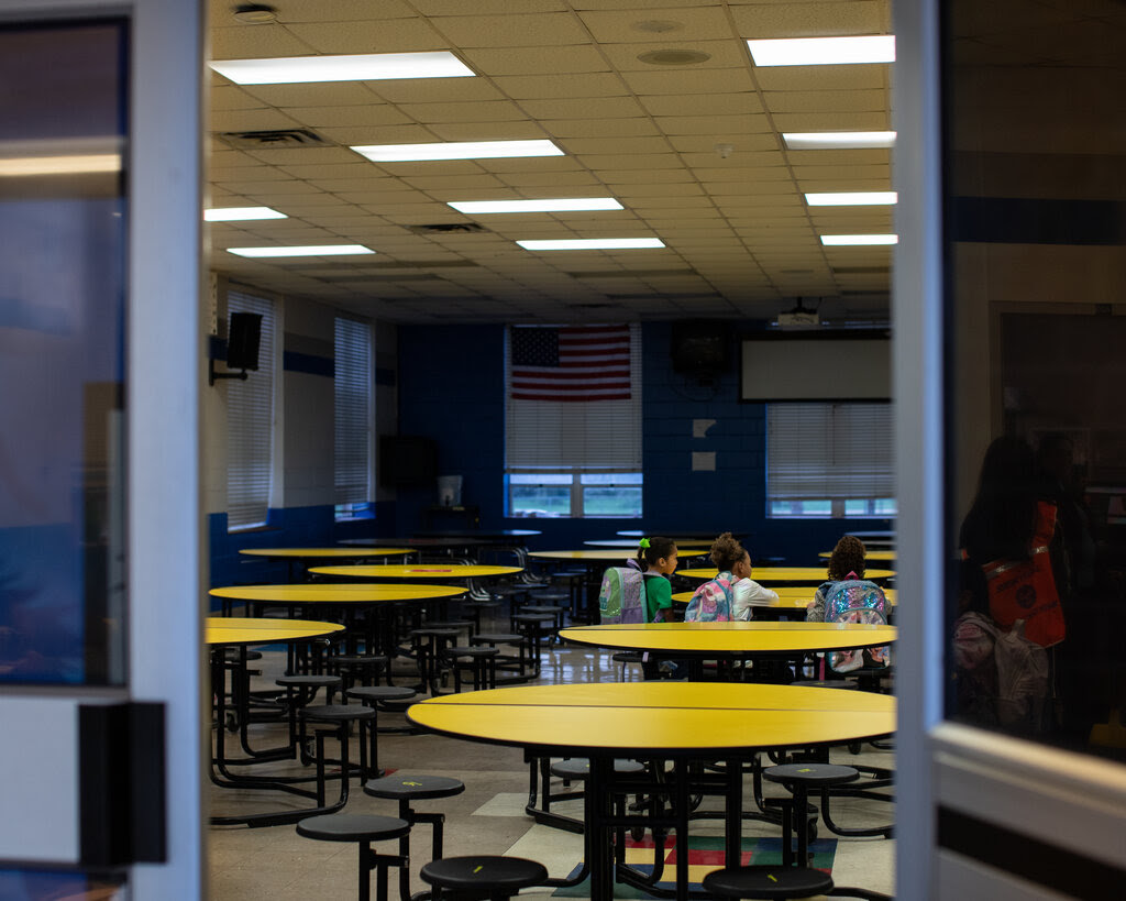 Three elementary-school students are seated around a circular yellow lunch table in a dining hall.