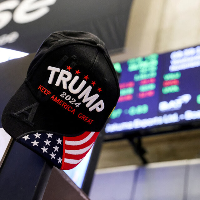 A Trump 2024 cap is seen in the foreground, with a New York Stock Exchange board blurred in the background.