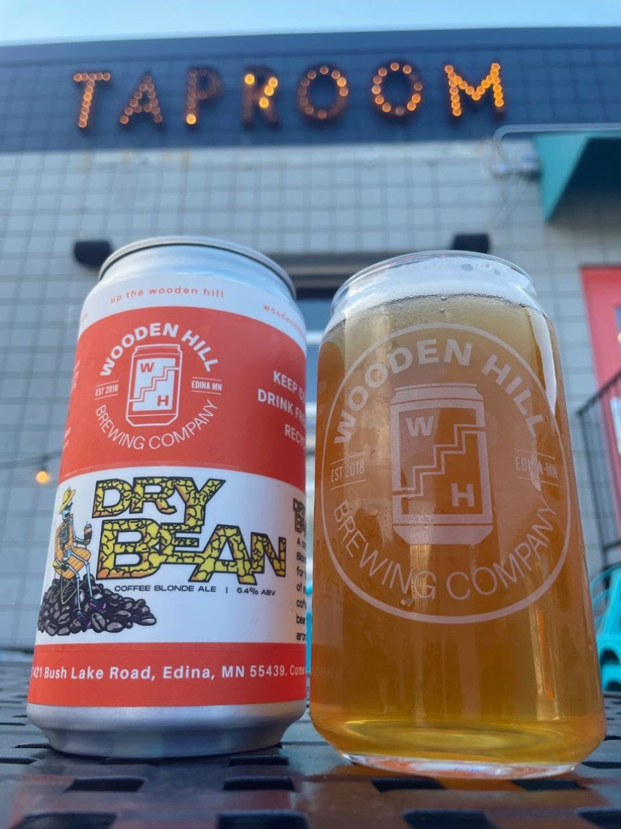 image of a crowler can of beer next to a pint glass filled with a light beer, with a light-up sign that says "TAPROOM" in the background.