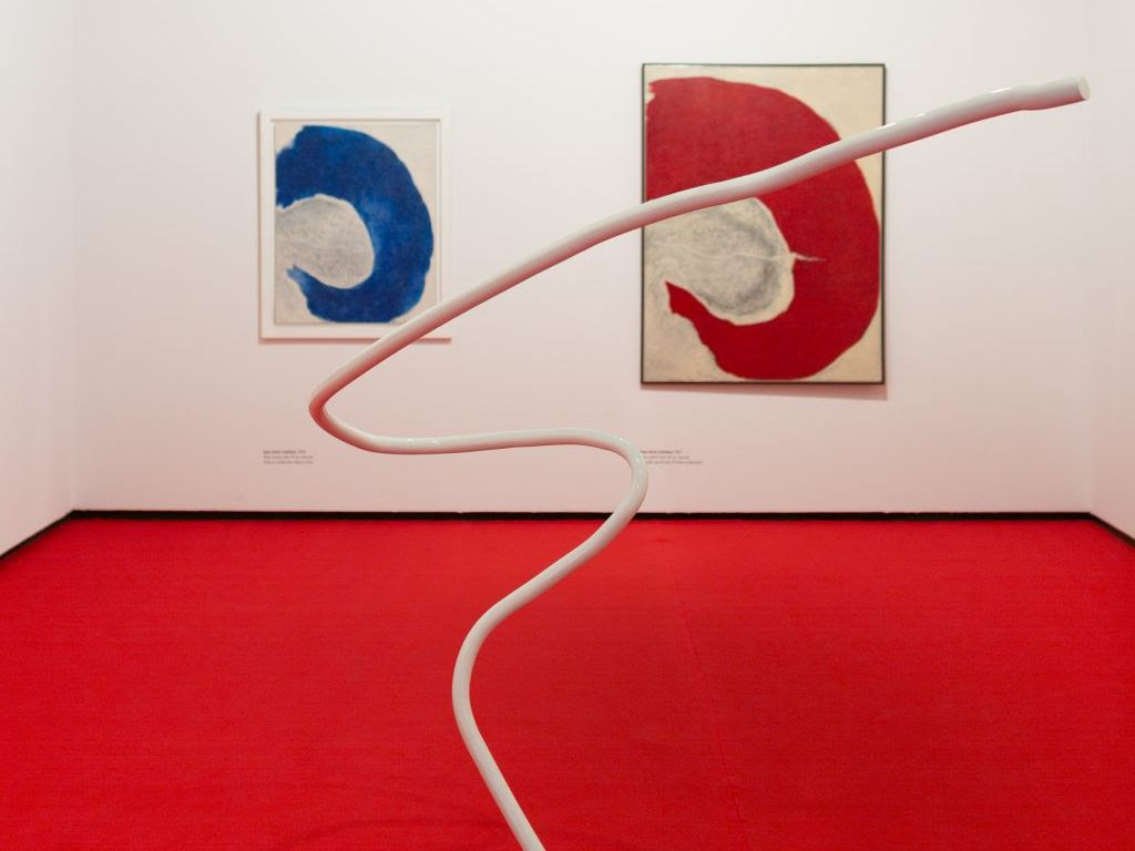 chão vermelho e parede branca ao fundo com duas pinturas. a pintura da esquerda é branca com uma mancha azul e a pintura da direita é branco com uma mancha vermelha. ao centro, uma escultura em tubo branco retorcido