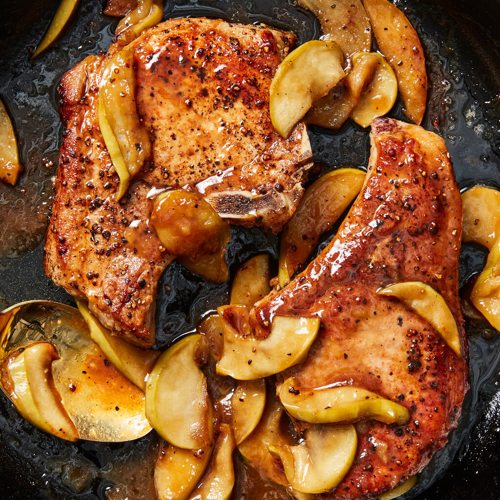 Two pork chops covered in a glaze with apple slices in a cast iron skillet.