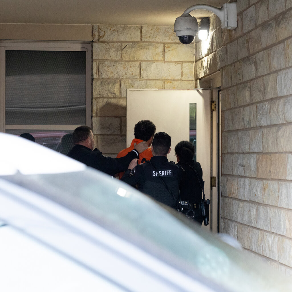 A group of police officers escort a man in an orange shirt to the door of a building.