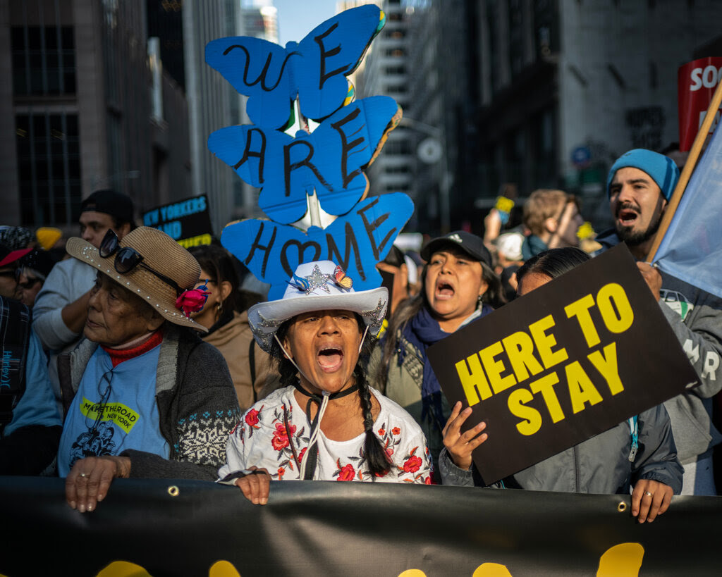 Several people marching in a protest as they hold a banner. One person holds up a sign that says, “Here To Stay.” Another sign reads, “We Are Home.” 