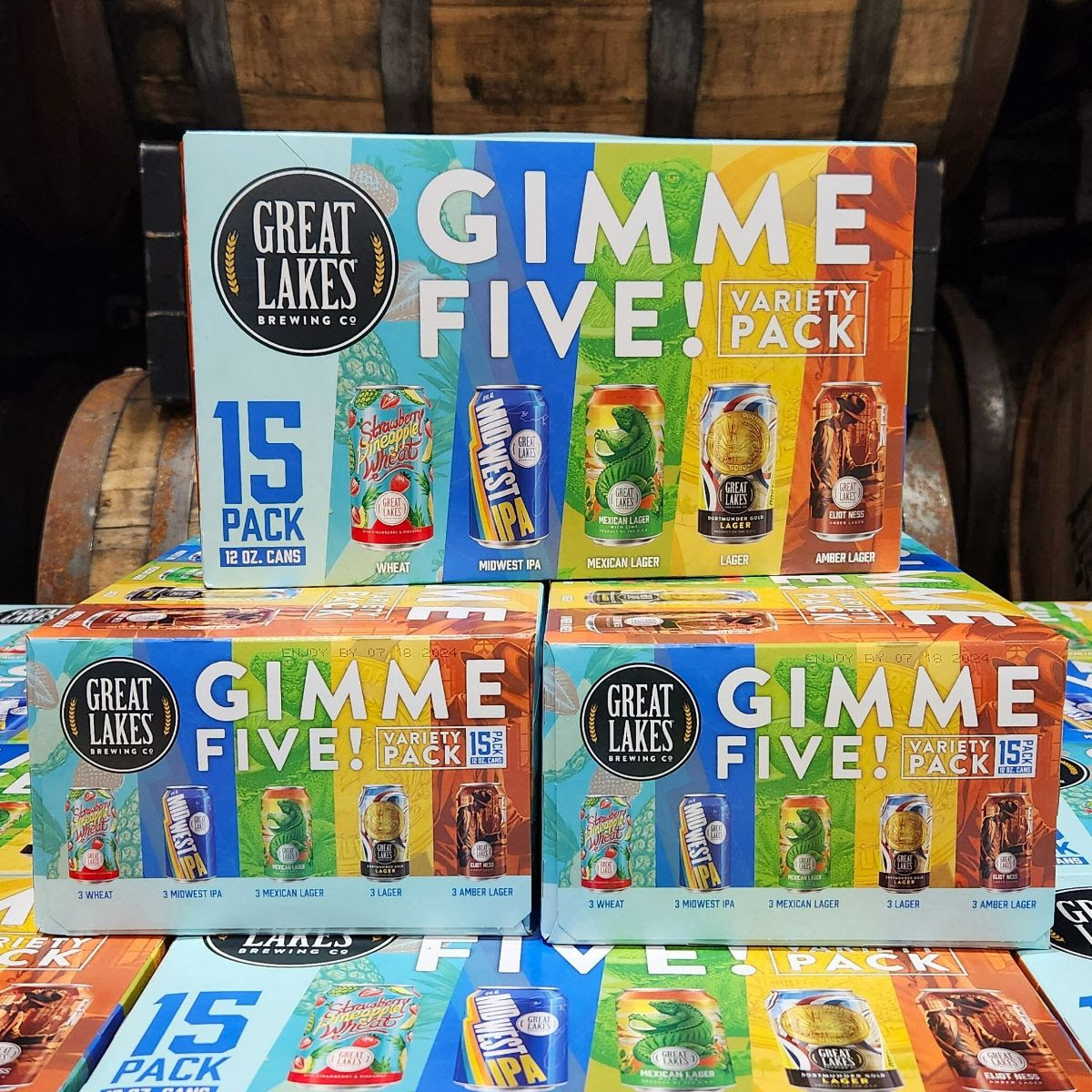 Many Summer Edition Gimme Five! Variety Packs stacked on each other in front of a set of wooden barrels.