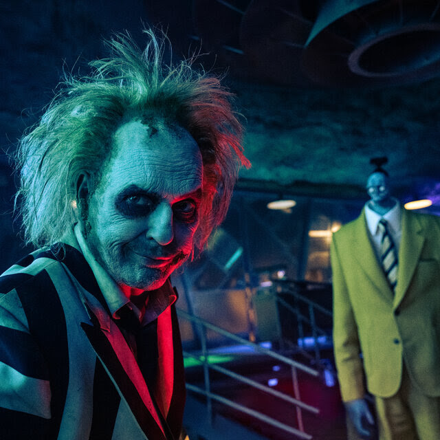 Michael Keaton as Beetlejuice, in a black-and-white striped suit with wild hair and white makeup on his face. 