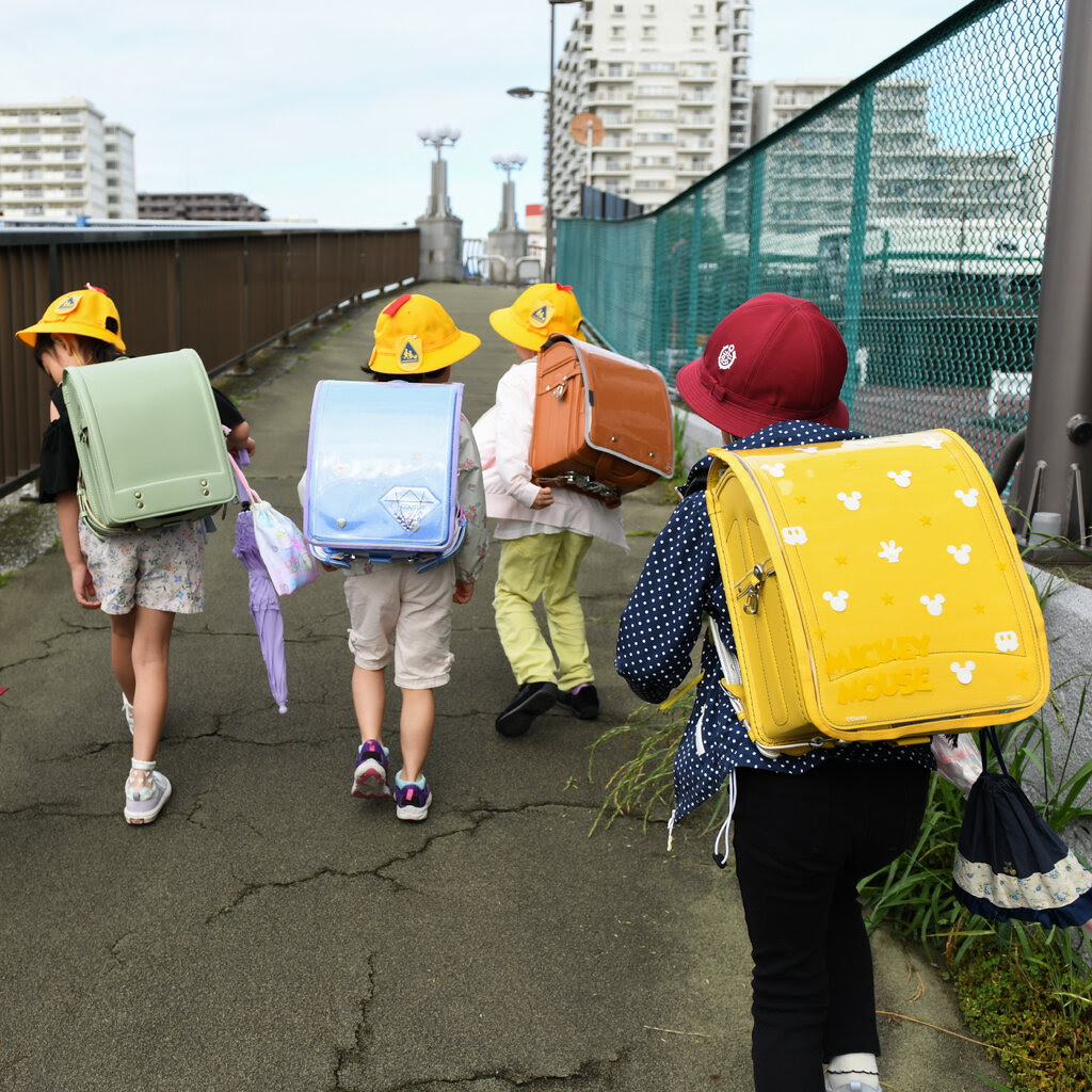 Five children, all wearing the same style of backpack but in different colors, walk down an elevated path in Tokyo. 