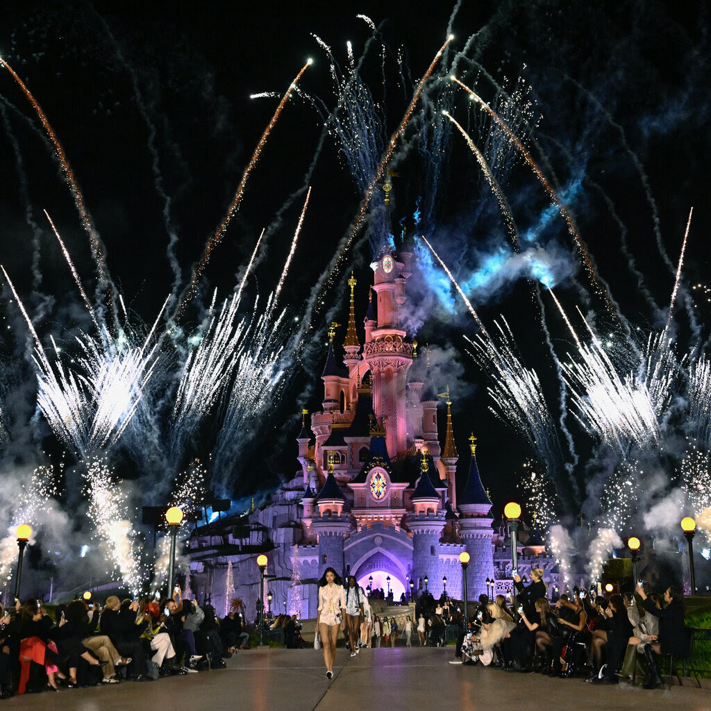 With the Sleeping Beauty Castle as a backdrop, and with fireworks exploding in the background, a line of models parades down a runway between seated showgoers.