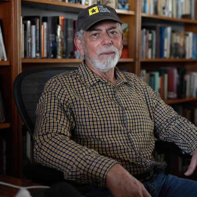 A man in a plaid shirt, bluejeans and baseball cap sits in front of shelves lined with books.