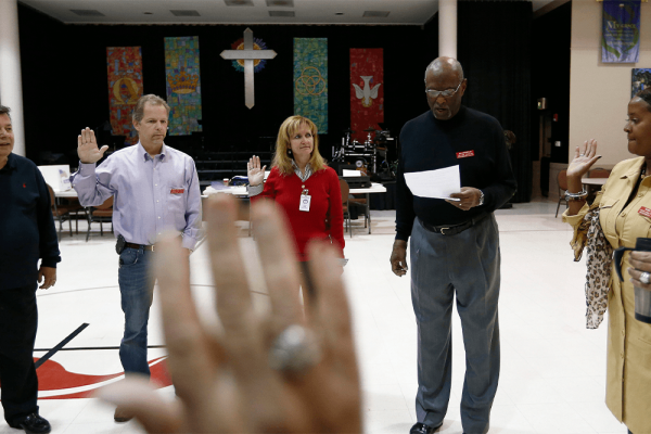 Multiracial group of adults stand with right hands held up in a church. 