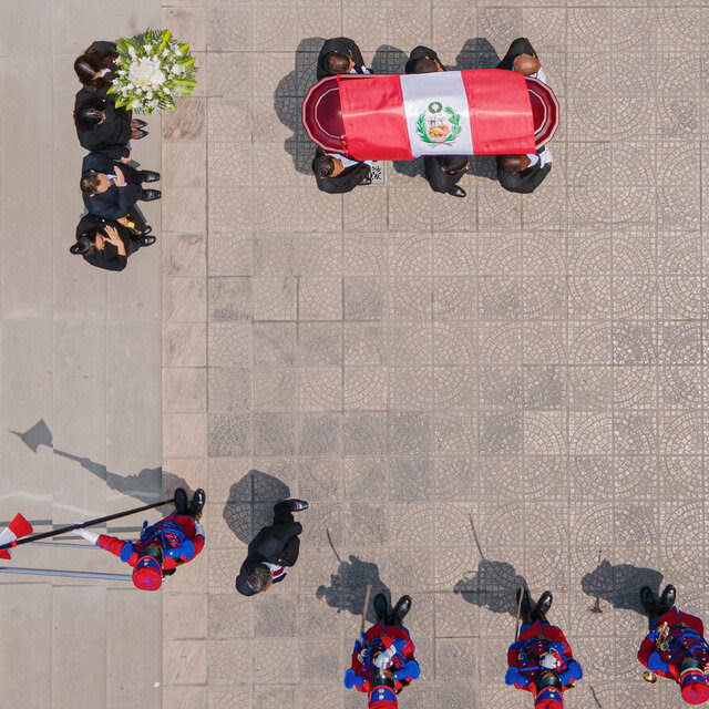 The coffin of Peru’s former president Alberto Fujimori, covered in a Peruvian flag, carried by six people arriving for his wake. Four people are standing behind the coffin, with one person holding a flower arrangement. A few uniformed musicians are off to the side.