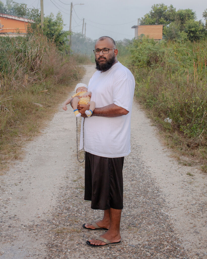 A portrait of Majid Khan, wearing a white T-shirt and black basketball shorts, holding a newborn baby on a gravel road with overgrown plants on either side.