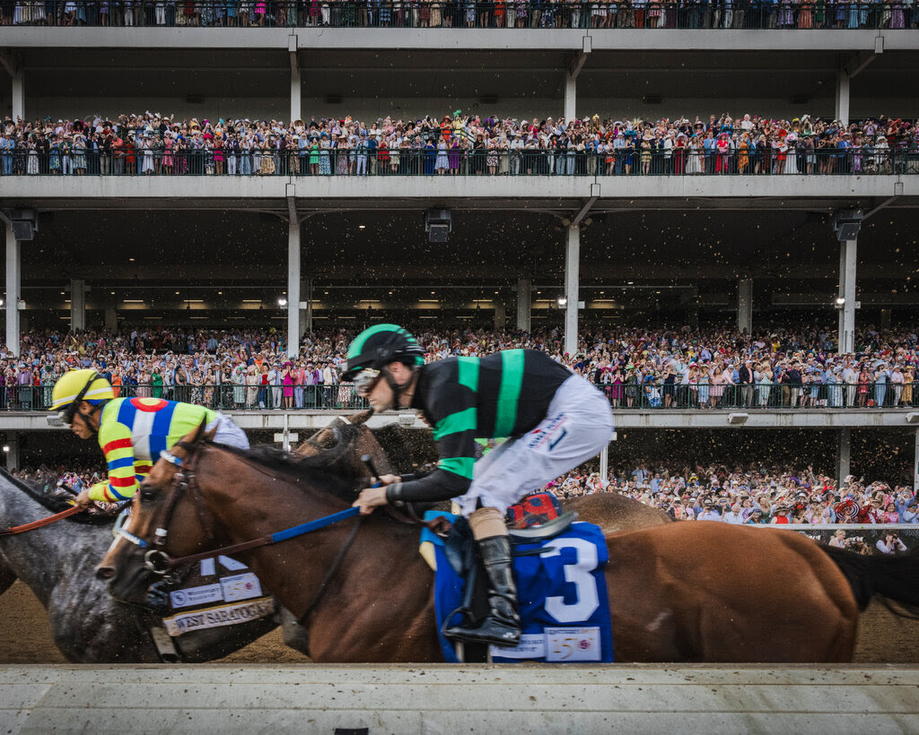 In the foreground, a jockey in black and green clothing rides a horse wearing the No. 3. A second horse, ridden by a jockey in multicolored clothing, is behind them. Crowded viewing stands are in the background.