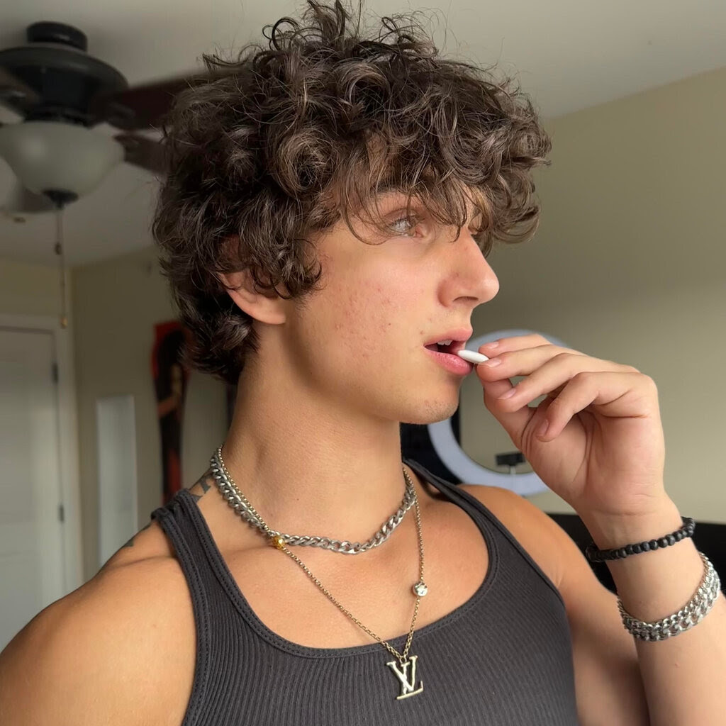 A young man with curly TikTok hair, a black tank top and a Louis Vuitton necklace stares pensively out a window as he holds a tablet of white gum up to his mouth.