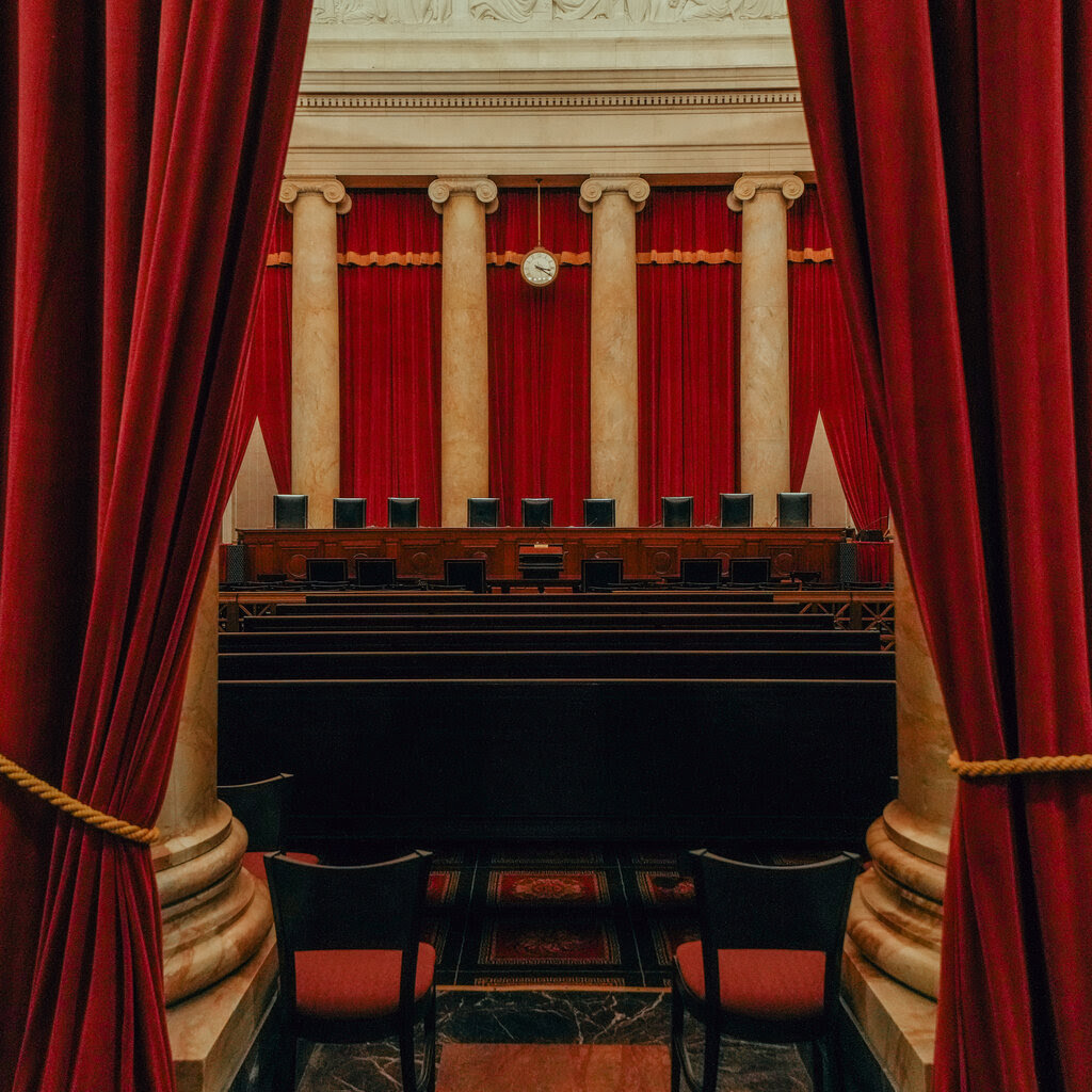 A view between two red curtains of benches. Nine leather chairs sit on a raised platform at the front of the room, with four columns behind.