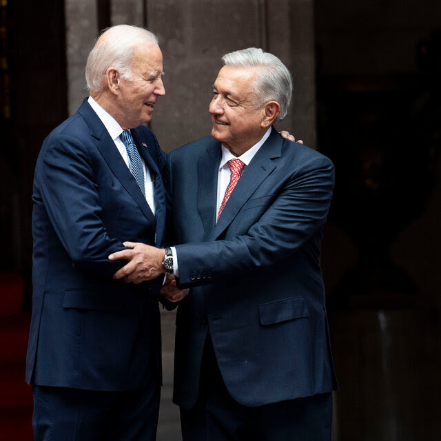 President Joe Biden and President Andrés Manuel López Obrador of Mexico City, both wearing dark suits, embrace next to an American flag.