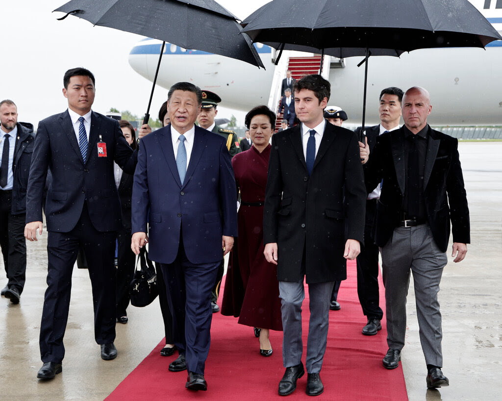 A group of mostly men walk down a red carpet from a plane. They are wearing suits. Xi Jinping is in front. 