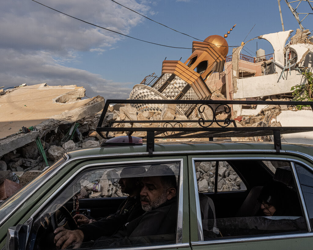 People in a car with a collapsed minaret and other rubble in the background. 