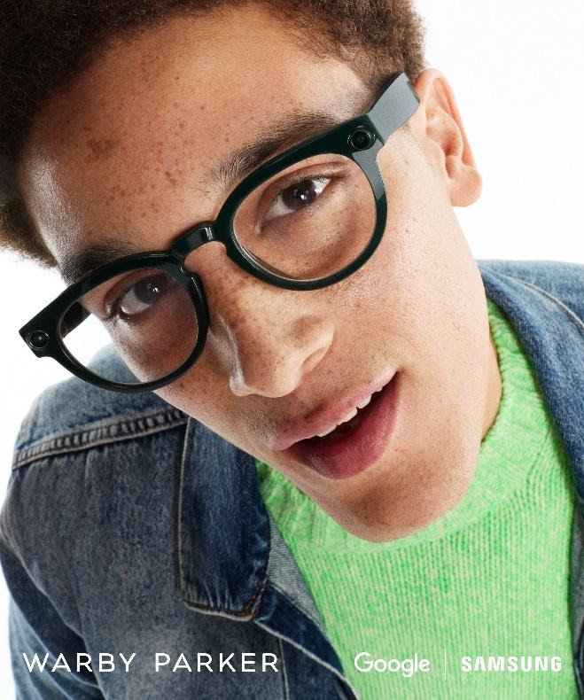 Persona joven con gafas modernas sonriendo, vestida con un suéter verde brillante y chaqueta de mezclilla, fondo blanco.
