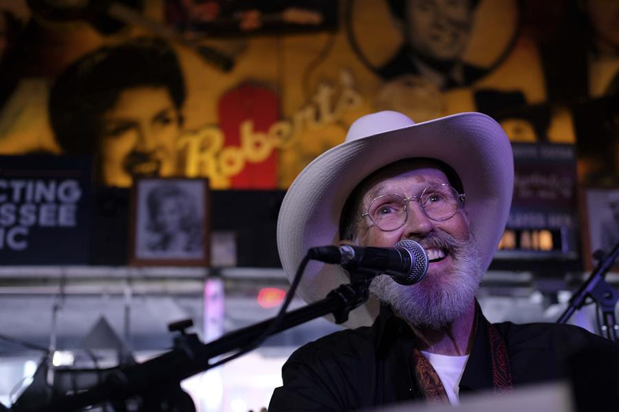 A man wearing a cowboy hat and glasses smiles while singing into a microphone.