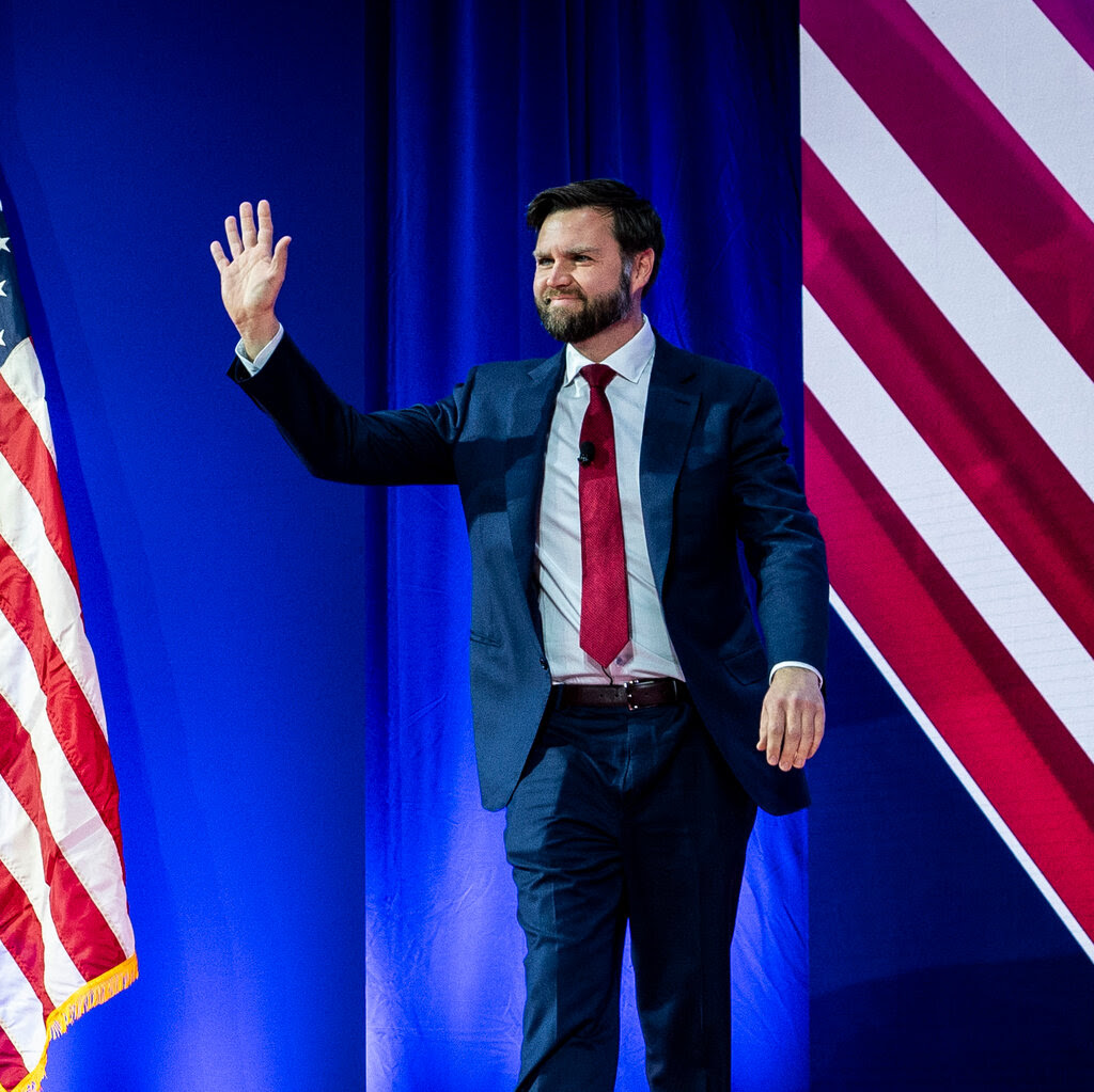 J.D. Vance in a blue suit and red tie waves beside an American flag.