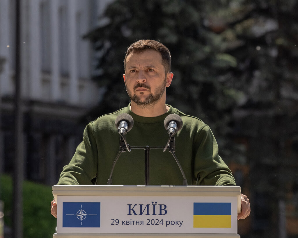 Volodymyr Zelensky, in a green top, speaking at a lectern outdoors.