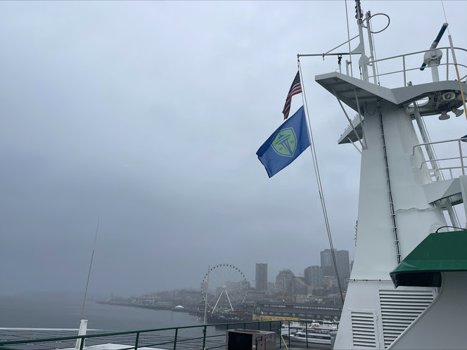 A ferry's mast with American and Sounders FC flags, against a the Seattle skyline with tall buildings and the Great Wheel wheel under a cloudy sky