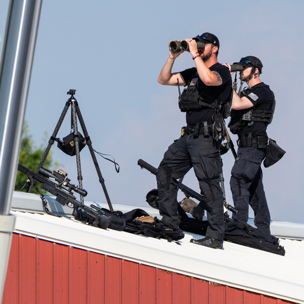 Two Secret Service snipers use binoculars to survey their surroundings from a white metal rooftop. A tripod and two long guns are lying on the rooftop at their feet.