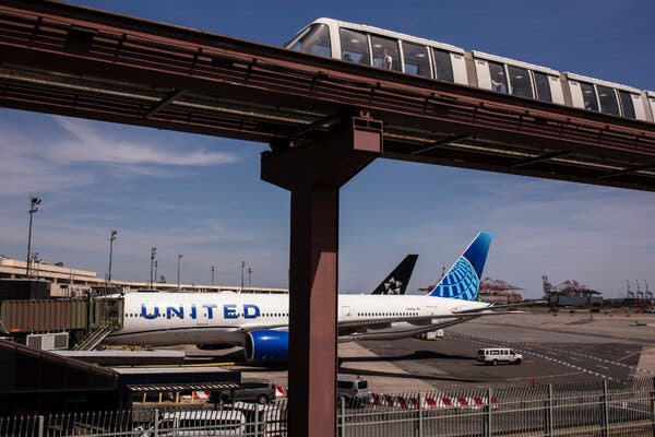 Un avión comercial blanco con “United” pintado en el lateral se ve en la pista de un aeropuerto. En primer plano, un tren lanzadera circula a toda velocidad por unas vías elevadas.