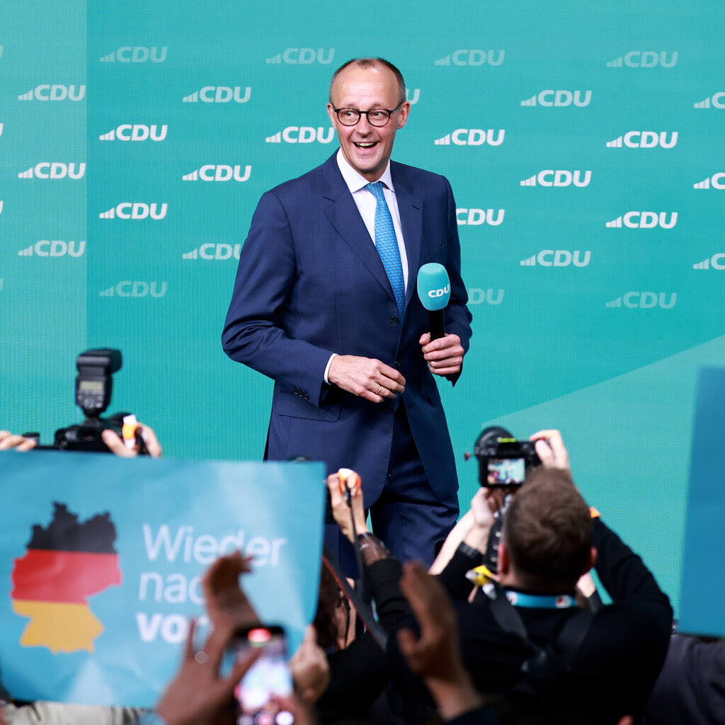 A man in a navy blue suit and sky blue tie walks across a stage with a microphone. In the foreground is a large crowd with people holding signs and cameras.