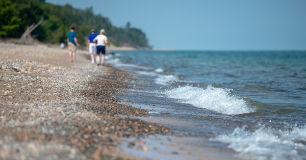 three people in shorts and T-shirts, off in distance, walk the sandy, pebbled Lake Superior shoreline as foamy waves roll in