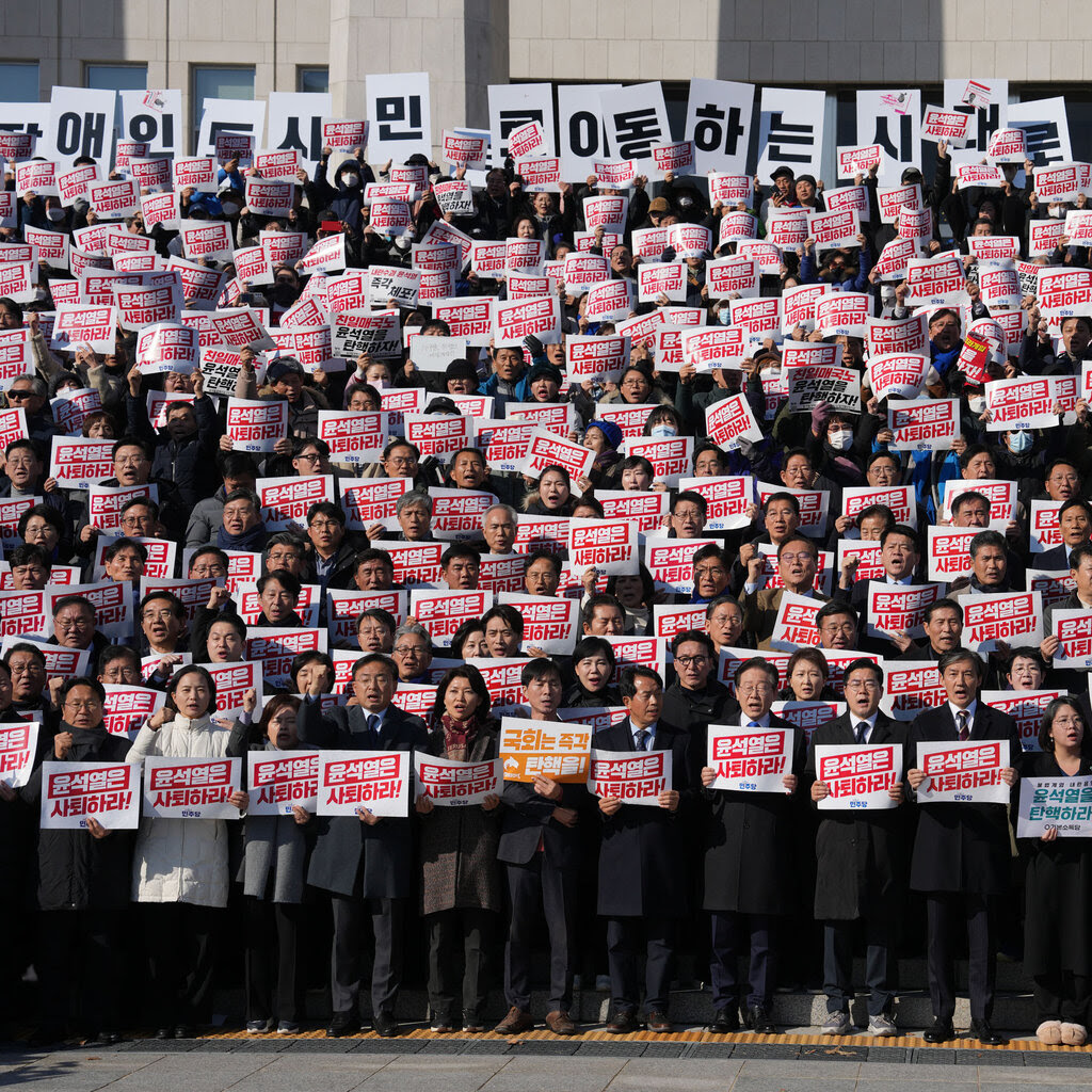 South Korean lawmakers protesting on the steps of the National Assembly holding white signs with red writing on them.