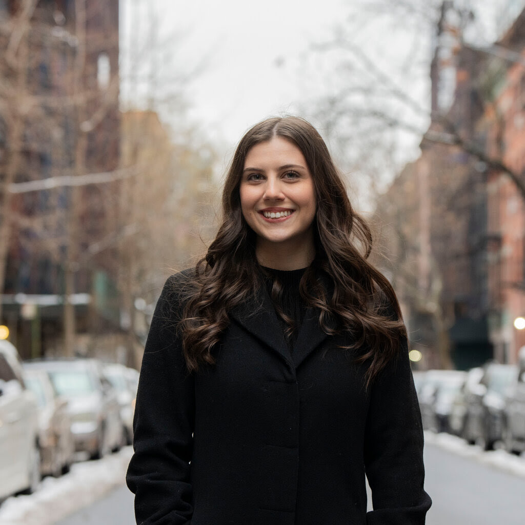 A woman with brown hair, wearing a black coat, smiles as she poses on a street.