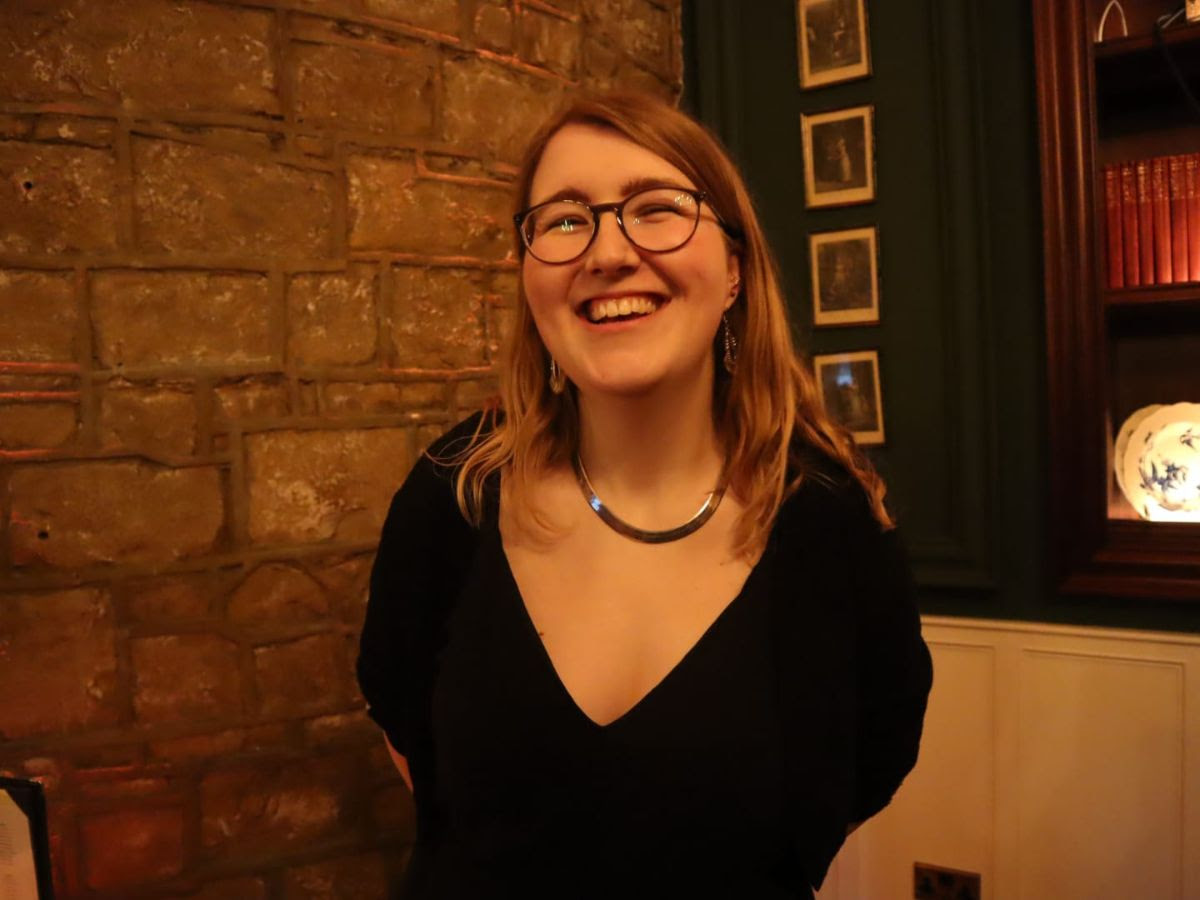 The writer Beth Storey, smiling in a black dress, stands in front of an exposed-brick wall