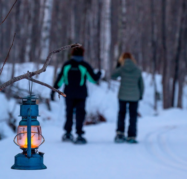 two people in winter gear and snowshoes on a forest trail, in the background, with a blue and glass, lit lantern in clear foreground focus