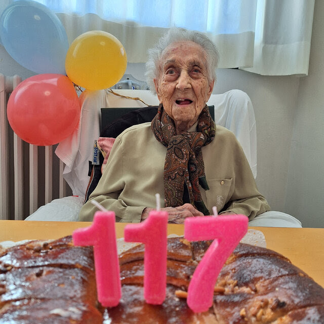 A woman sits in a chair with two balloons tied to it, in front of a cake with three candles in the shape of the number 117. 