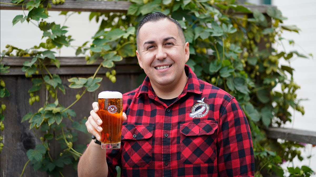 North Coast Brewing Assistant Brewmaster Obed Salazar stands in front of a fence covered in hop vines, holding a pint of Wet Coast Pale Ale