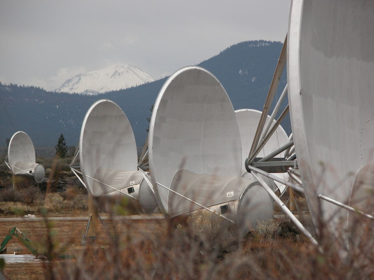 Several large satellite dishes in a field, with snowy mountains in the background.