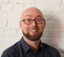 A middle-aged Caucasian bald man with black rimmed glasses and beard pictured to the shoulder with a dark button down shirt against a white brick wall.