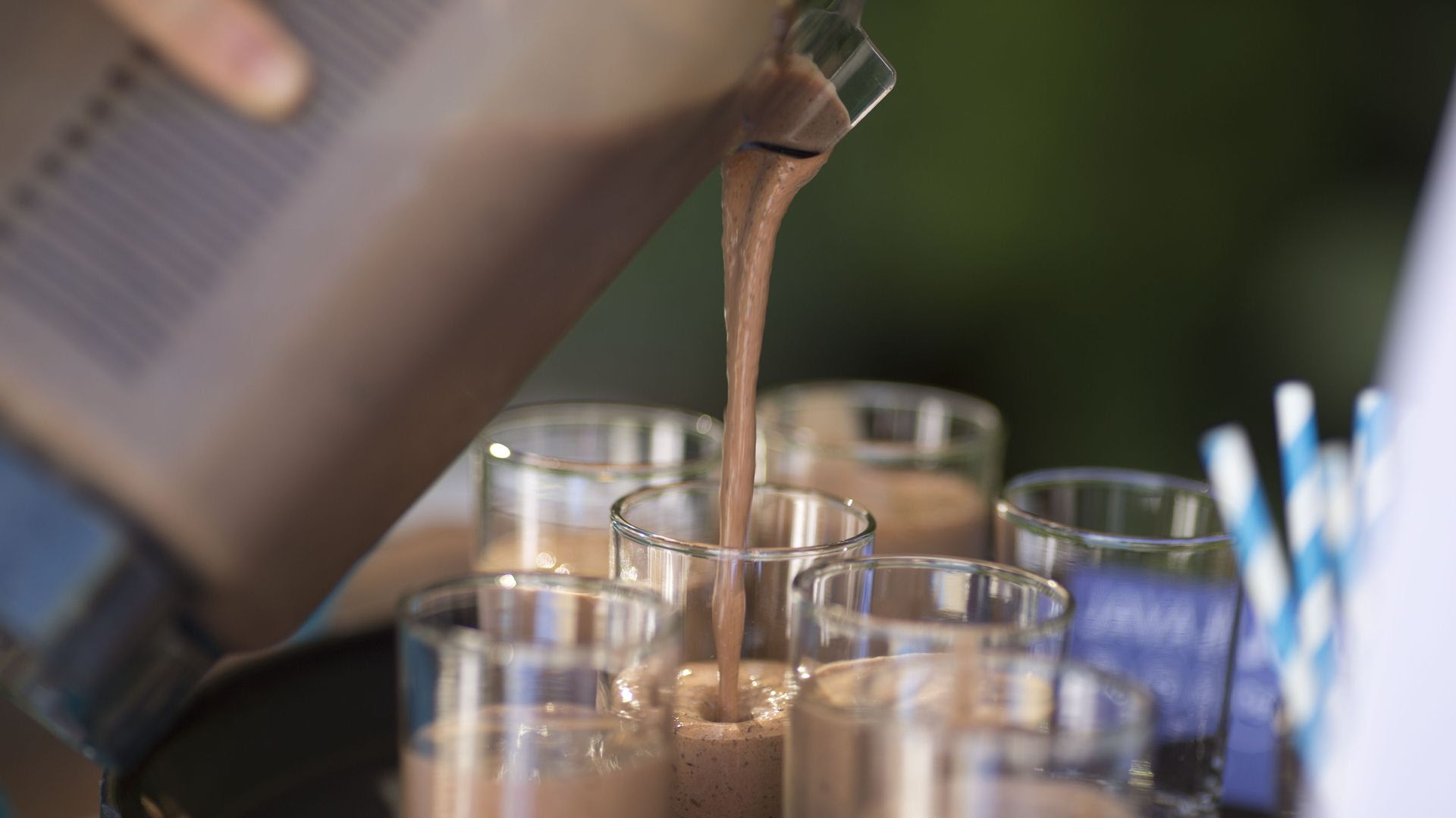 An image of six glasses on a tray with someone pouring a chocolate protein shake into them